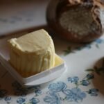 Soft butter in a small white butter dish on a floral tablecloth beside a loaf of bread