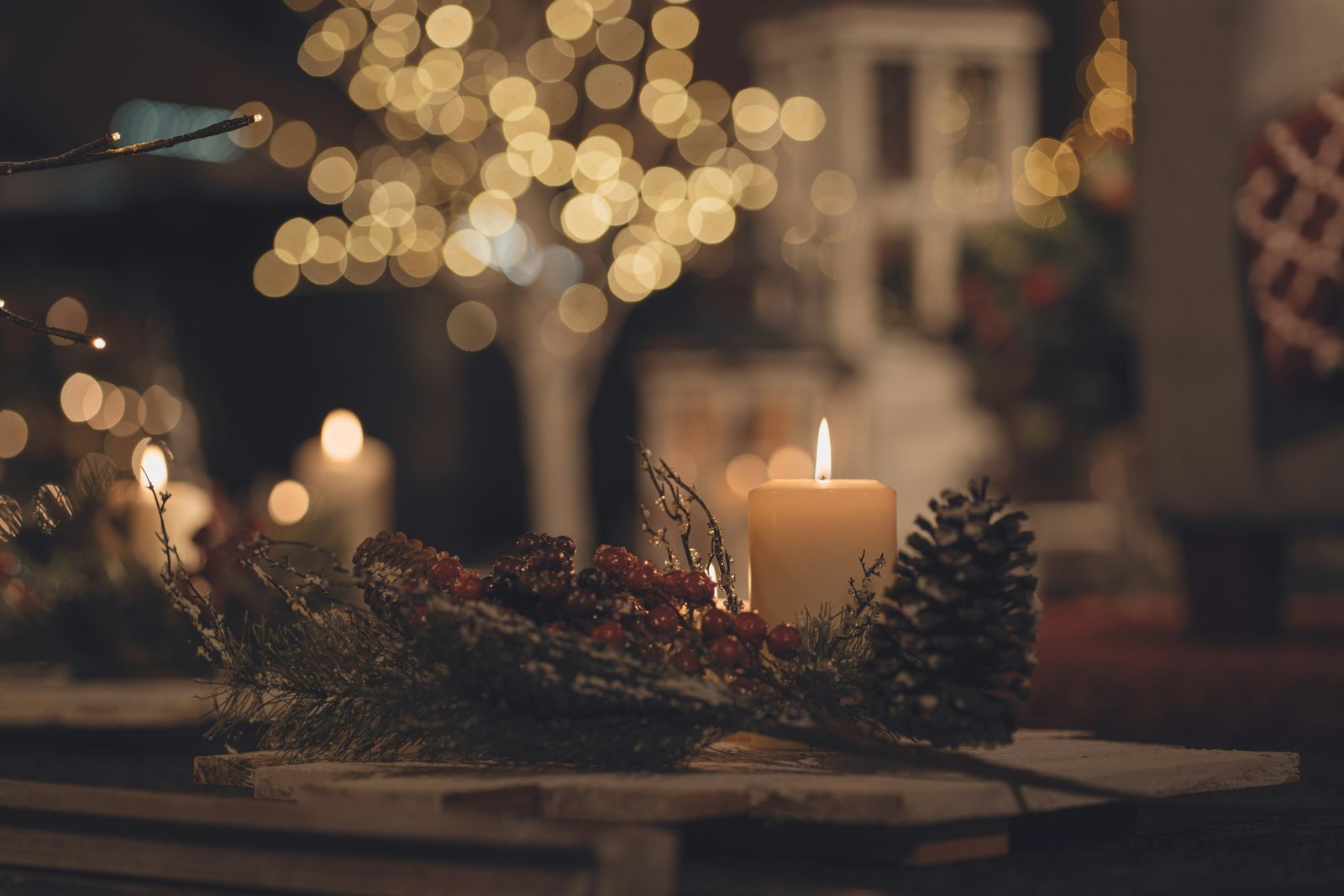 Best Christmas candles: a lit pillar candle with pine cone, berries and fir sprigs in front of glowing holiday lights