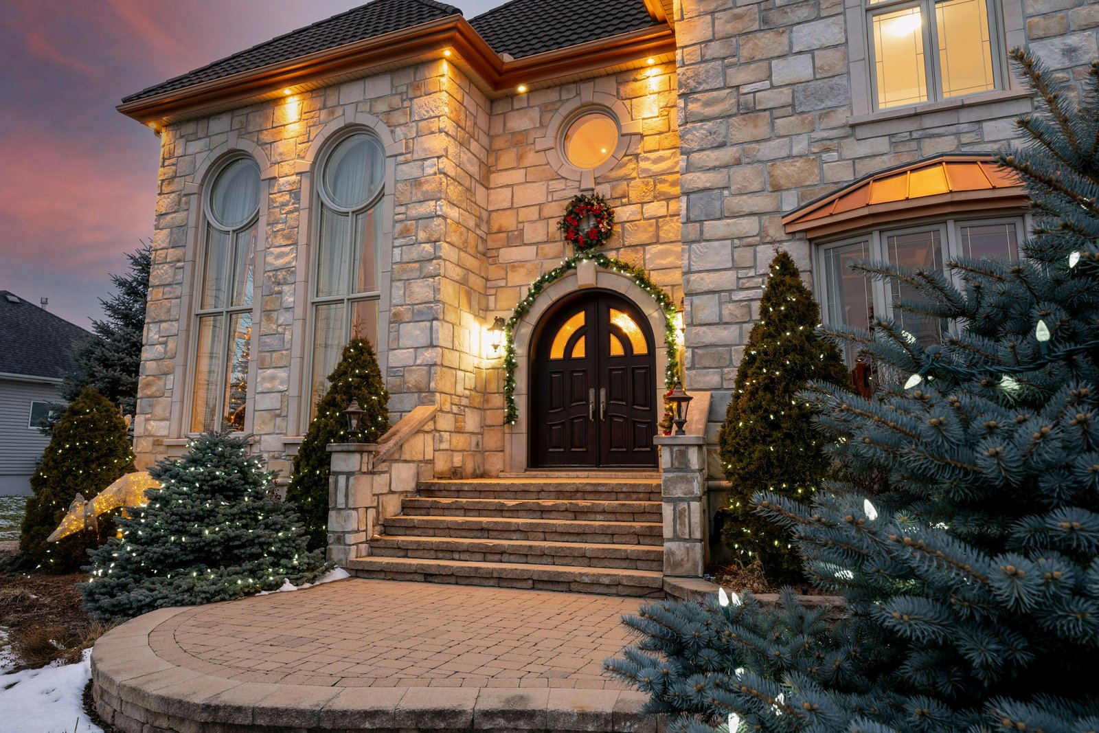 Christmas doormat ideas: a stone house entry with double doors, a wreath, garland and lit trees by the front steps
