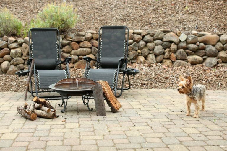 Best fire pits for a backyard patio: a steel fire pit bowl with two lounge chairs and stacked firewood on brick pavers