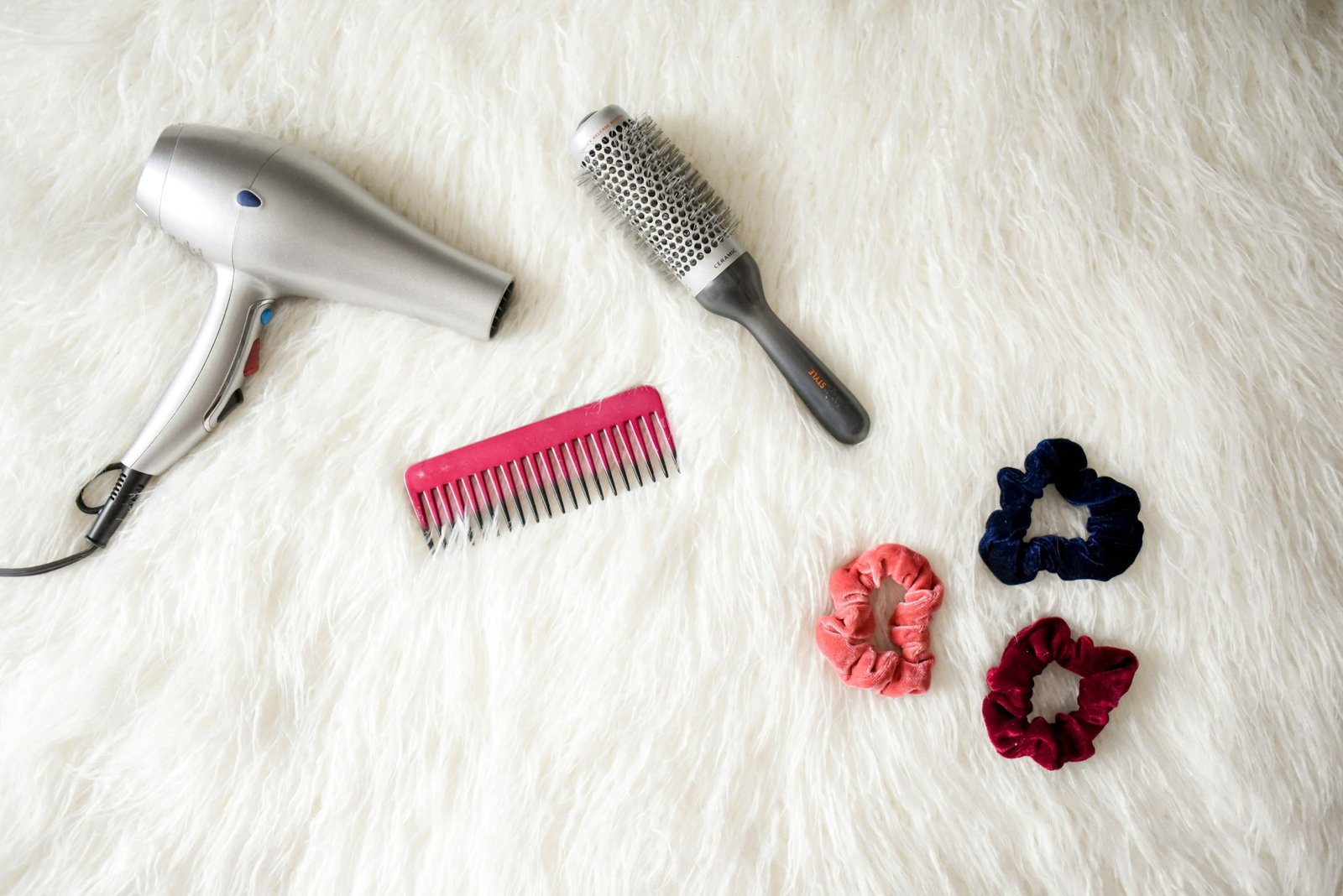 Hair styling tools flat lay: a hair dryer, round brush, wide-tooth comb and velvet scrunchies on a white rug