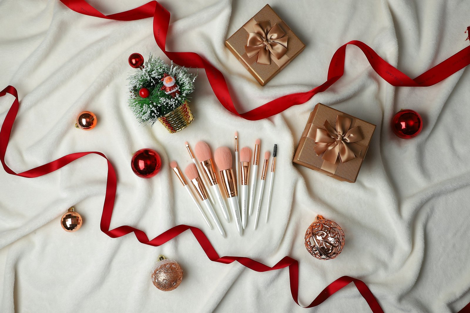 Luxury beauty advent calendar gift idea: makeup brushes, gold gift boxes and red ribbon on a white throw