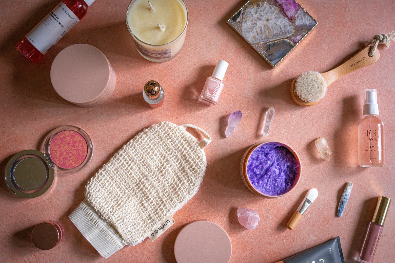 How to use a body scrub: exfoliating mitt, body brush and skincare jars laid out on a pink surface