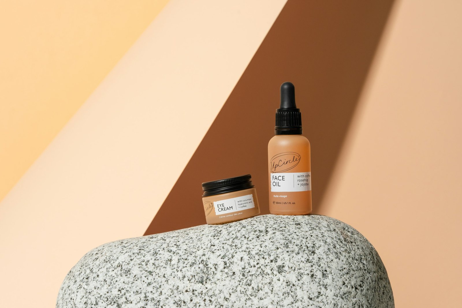 Best luxury eye cream: an amber eye cream jar and a face oil bottle on a speckled stone against a peach backdrop