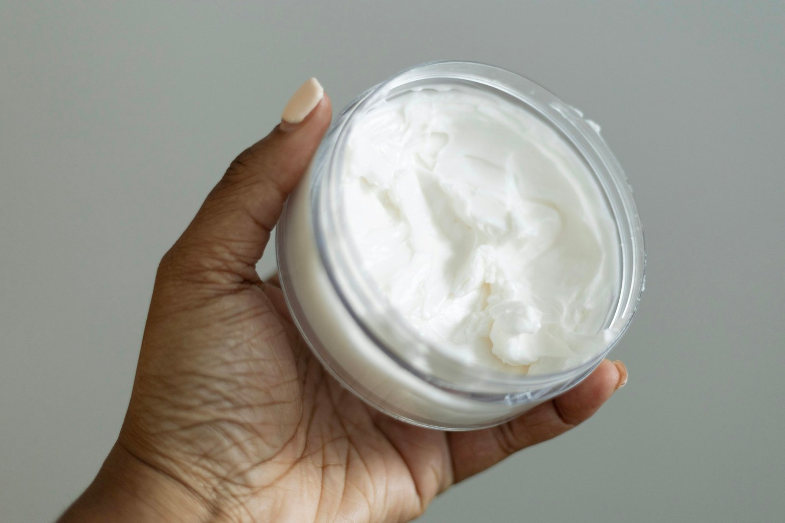 Best luxury face moisturizer: a hand holding an open jar of rich white face cream against a soft gray wall