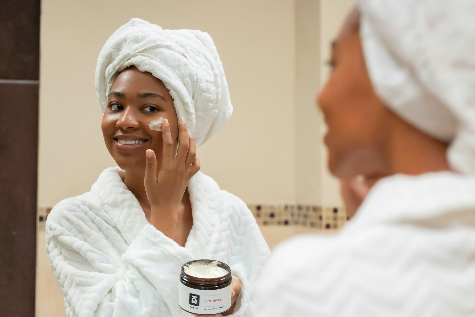 Luxury face cream being applied: a woman in a white robe and towel dots moisturizer on her cheek in a bathroom mirror