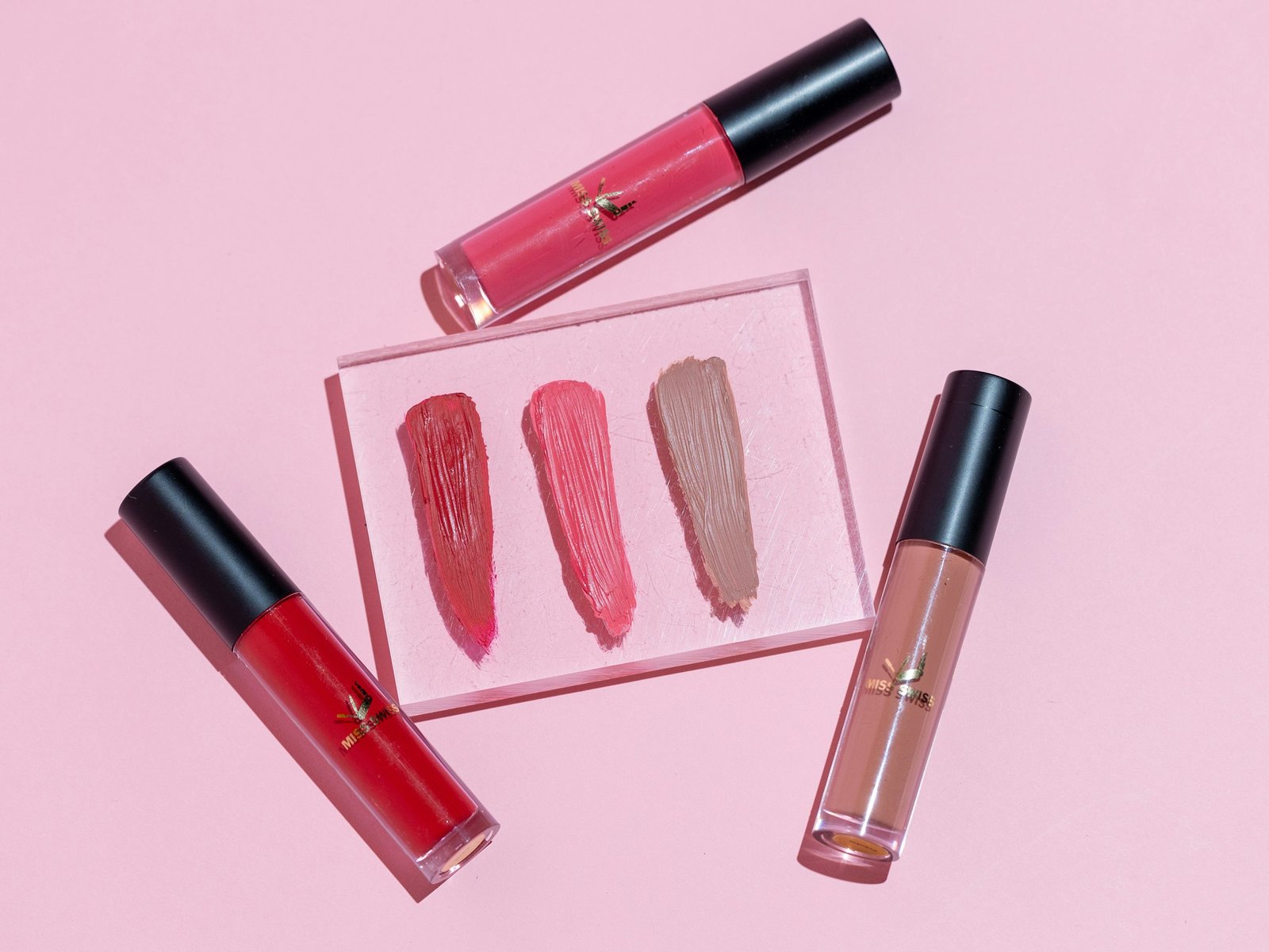 Luxury lip gloss and liquid lipstick swatches in red, pink and nude on a clear block, showing gloss vs creamy finish