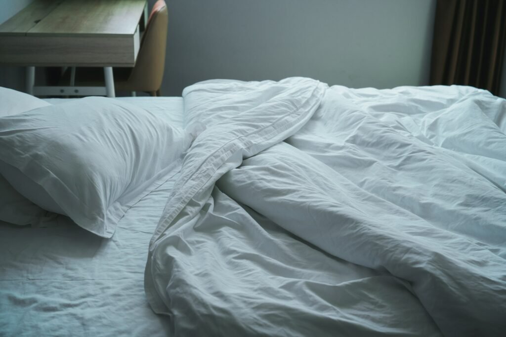 Unmade bed with white sheets ready for laundry day, part of a mattress protector vs mattress pad care routine
