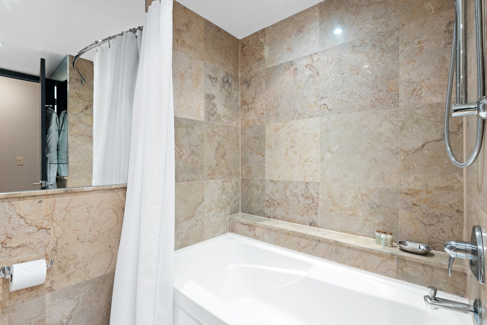 Best shower curtains: a white fabric shower curtain hung high on a curved rod over a bathtub in a stone-tiled hotel-style bathroom