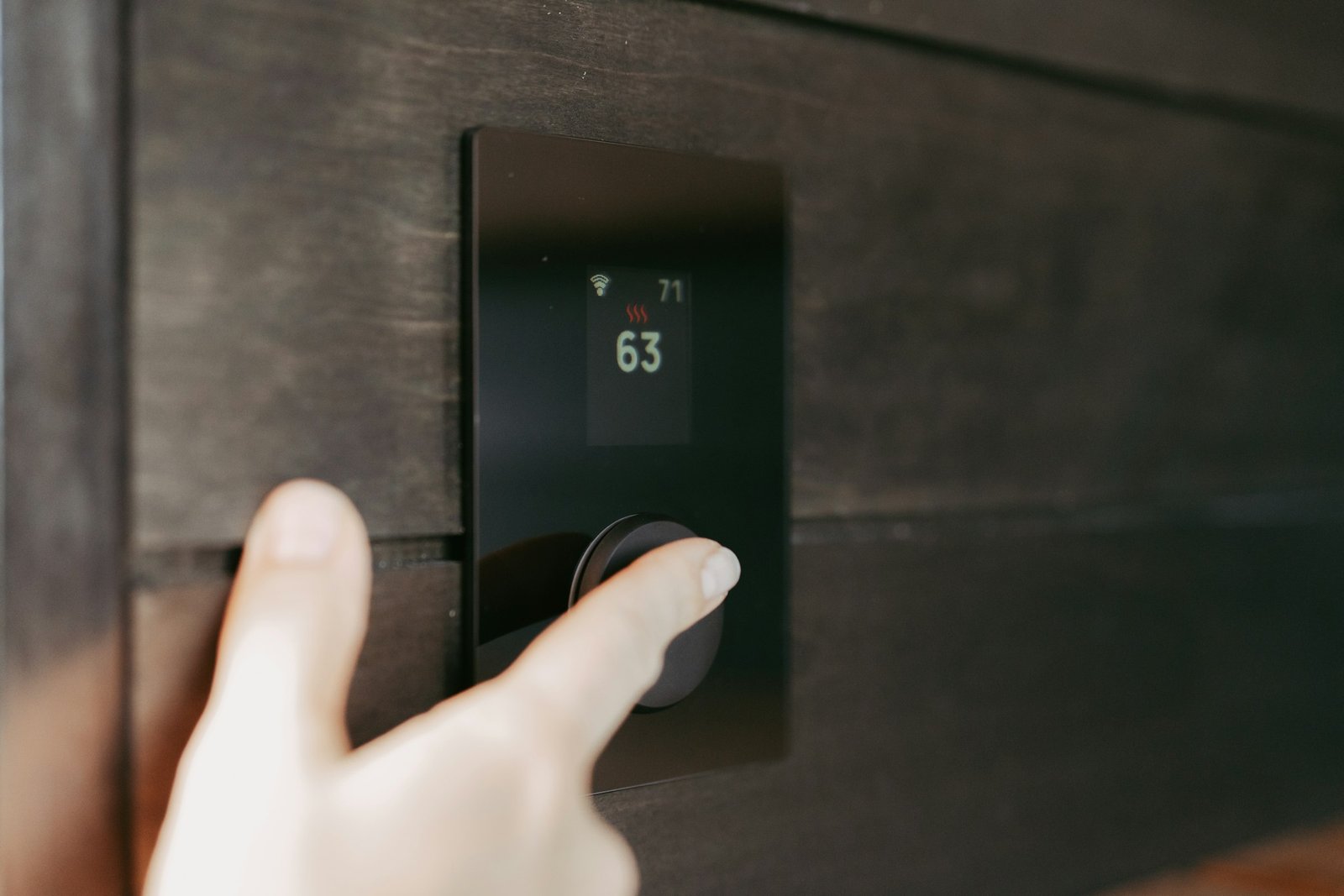 Best smart thermostat for heating savings: a hand turning the dial of a smart thermostat set to 63 degrees on a dark wood wall