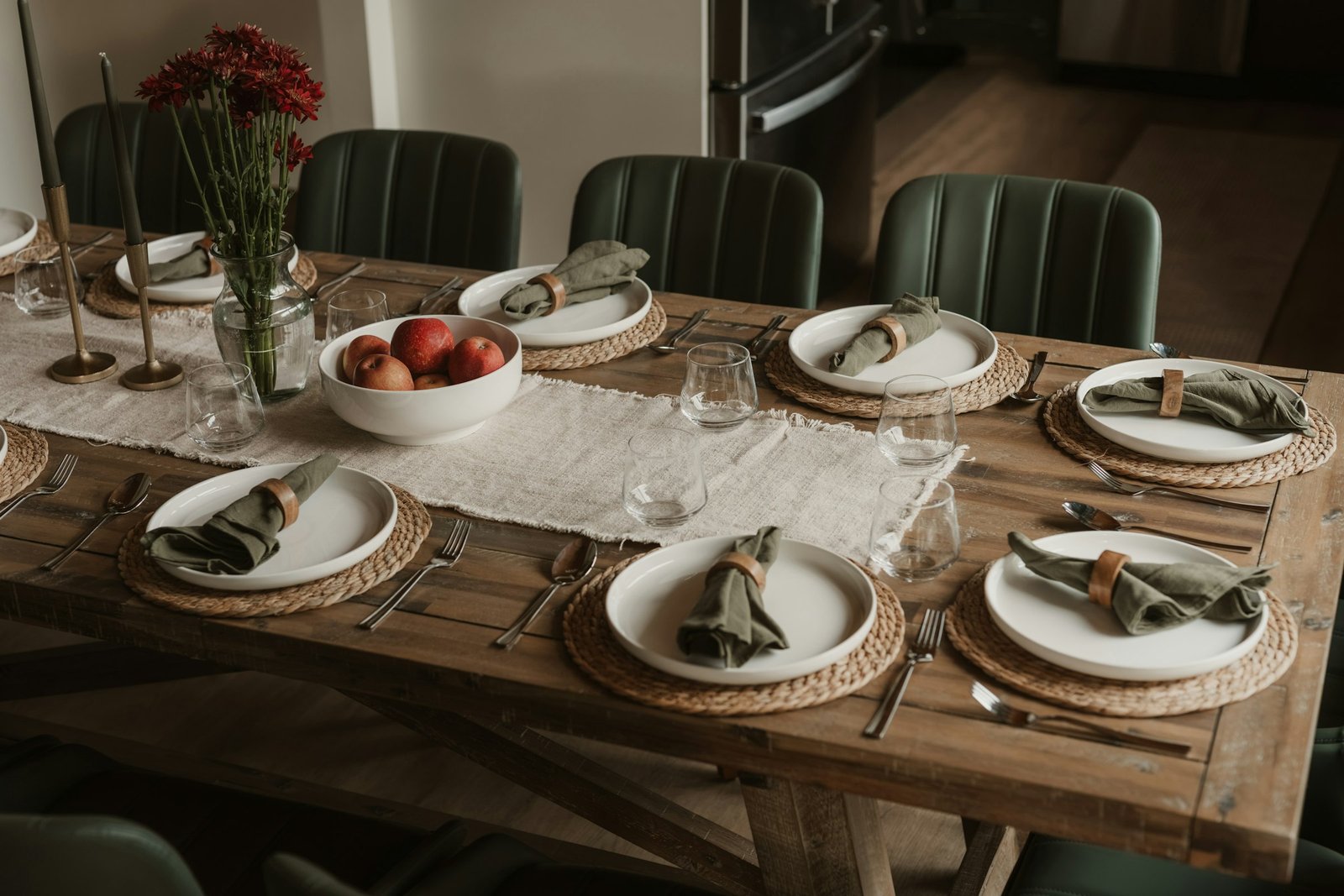 Thanksgiving table decor: wooden dining table with a white runner, sage napkins on woven chargers, taper candles and a low bowl of apples