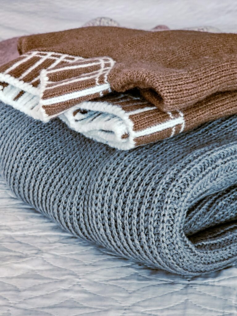 Folded knit sweaters and a blanket on a bed, ready to pack into under bed storage containers with lids