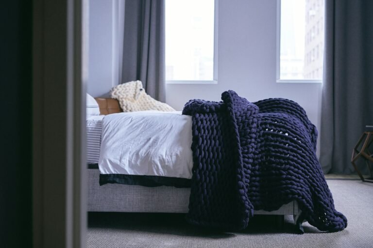 Best weighted blanket: a navy chunky knit weighted blanket draped over a white bed by tall windows