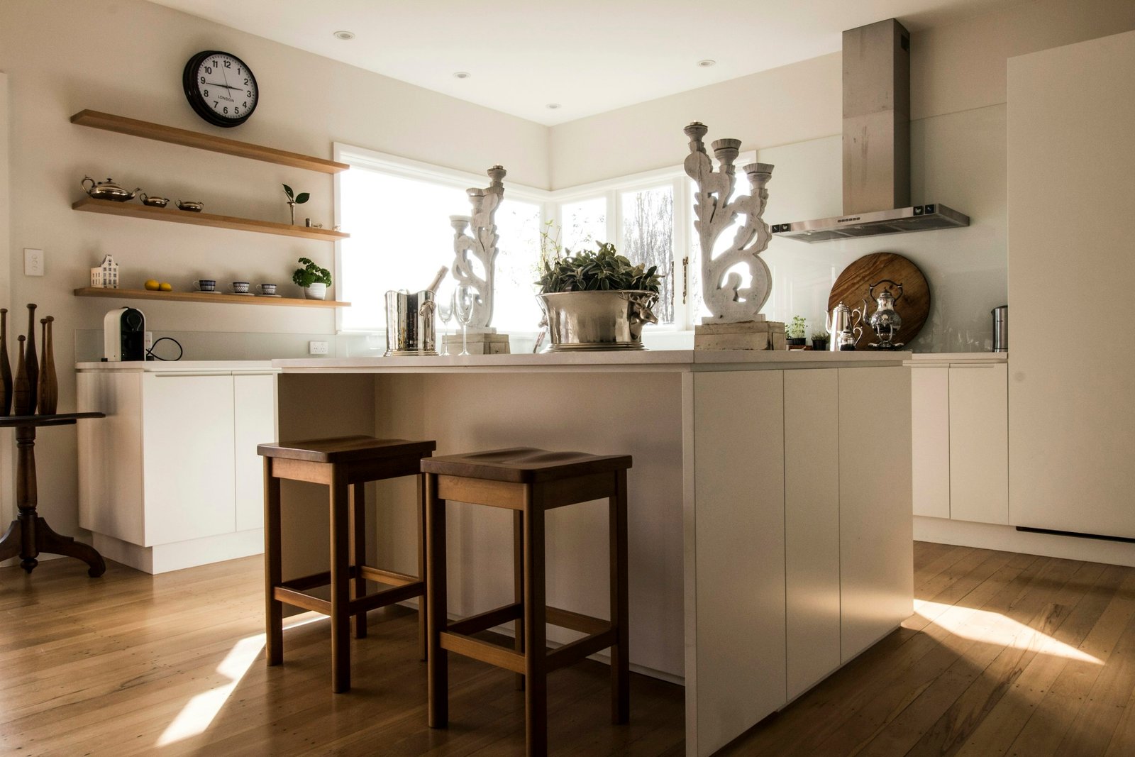 Black kitchen wall clock above open wooden shelves in a bright white kitchen
