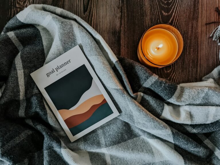 Cozy home gifts: a plaid throw blanket, a lit candle and a planner on a wood table