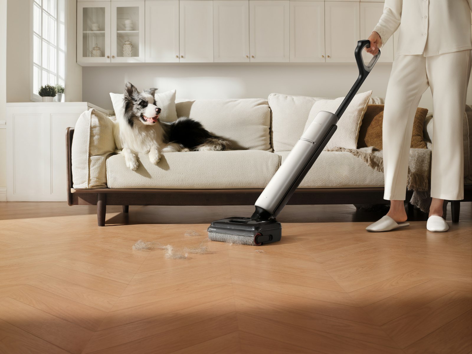 Dyson V15 vs V8: a cordless stick vacuum picking up pet hair on a light wood floor while a dog rests on the sofa