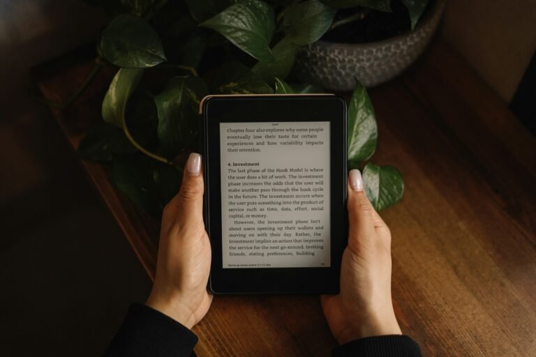 Kindle Paperwhite vs Colorsoft: hands holding a Kindle e-reader over a wooden table with a potted plant