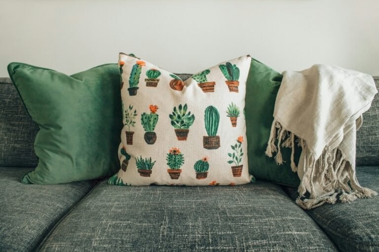 Olive green throw pillows on a textured gray sofa with a cactus print pillow and a cream fringed throw