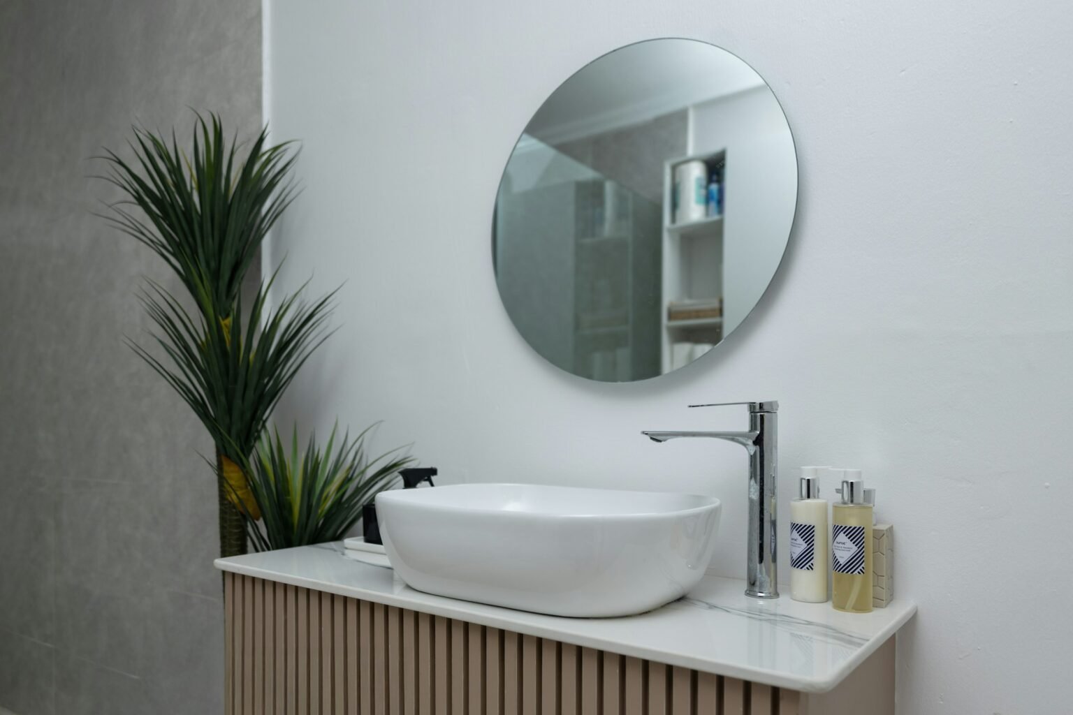 Round bathroom mirror ideas: a frameless circle mirror above a vessel sink with a plant